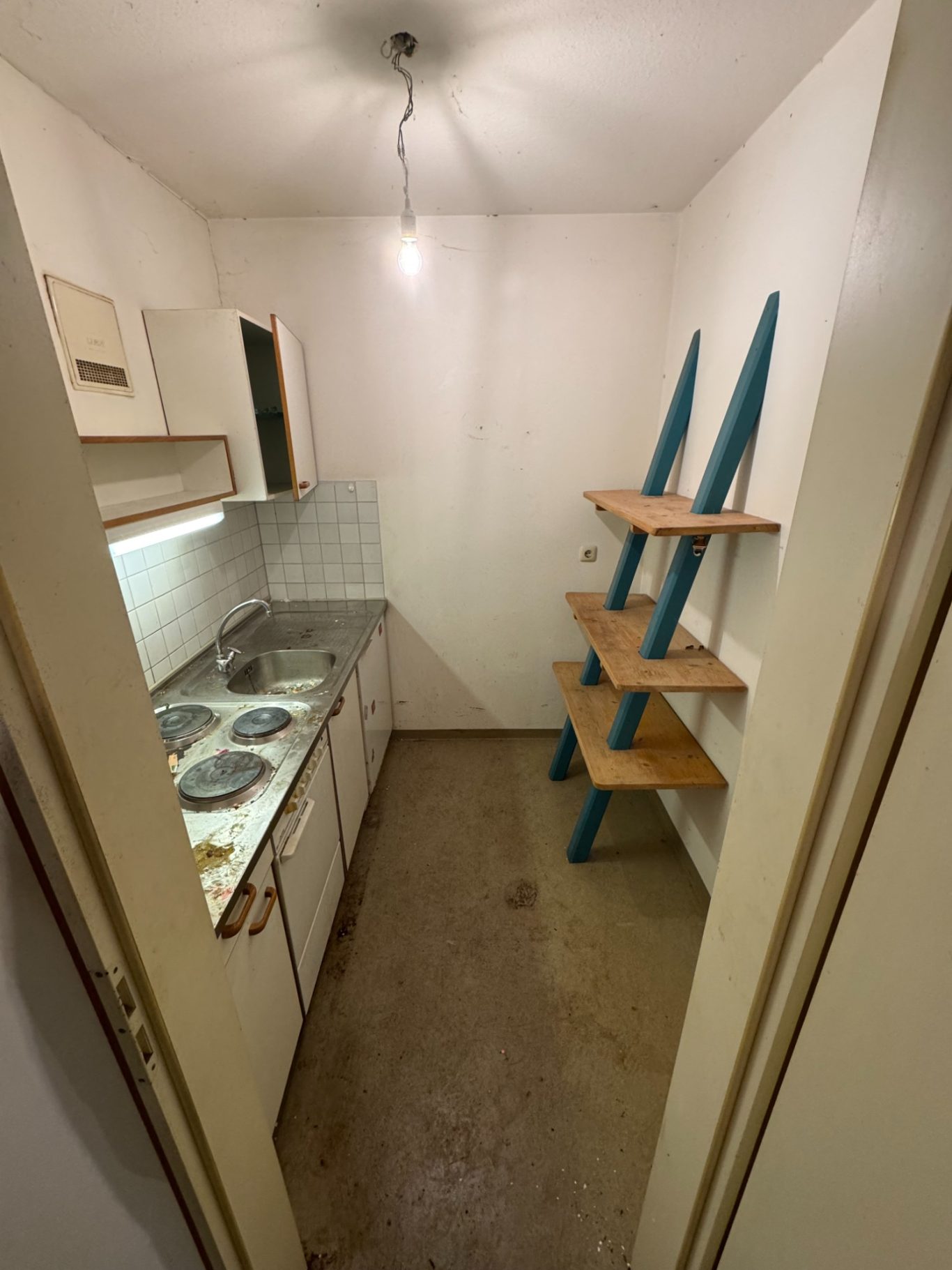 Kleine Küche mit weißen Schränken, Kochplatten und einem Holzregal an der Wand.